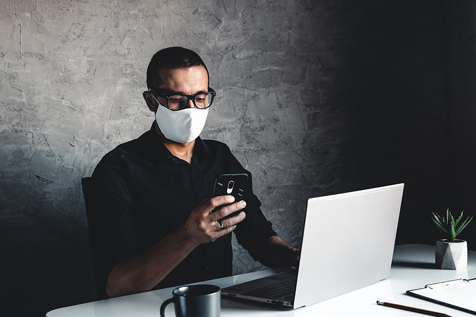 masked man at a computer holds a smart phone